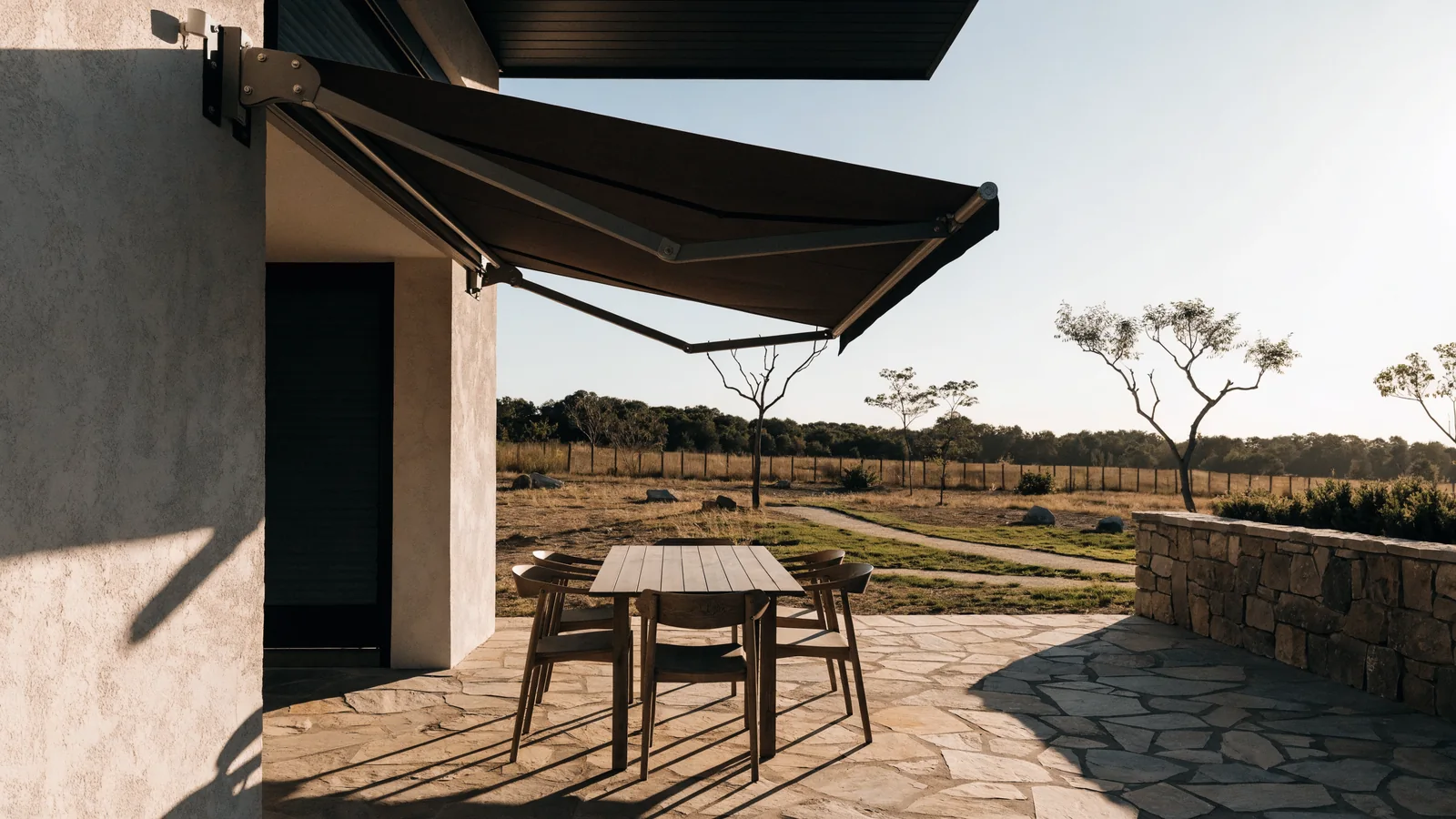 A folding-arm awning fully extended on its articulated arms over a stone-paved covered patio of a modern Midstream estate home, a small wind sensor on the wall bracket, a young landscaped estate park and wide open Highveld sky beyond