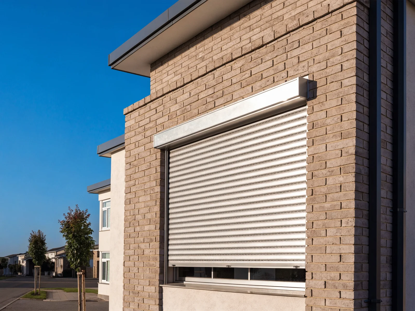An aluminium roller shutter of rigid horizontal slats rolling down from a slim headbox on the outside of a large ground-floor window of a modern double-storey Midstream estate home, under a wide open Highveld sky with young ornamental street trees in front